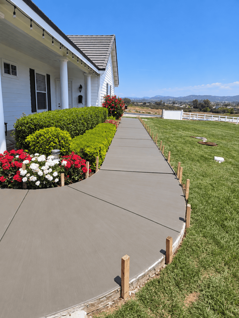 Newly poured concrete sidewalk with wooden stakes in front of a white house.