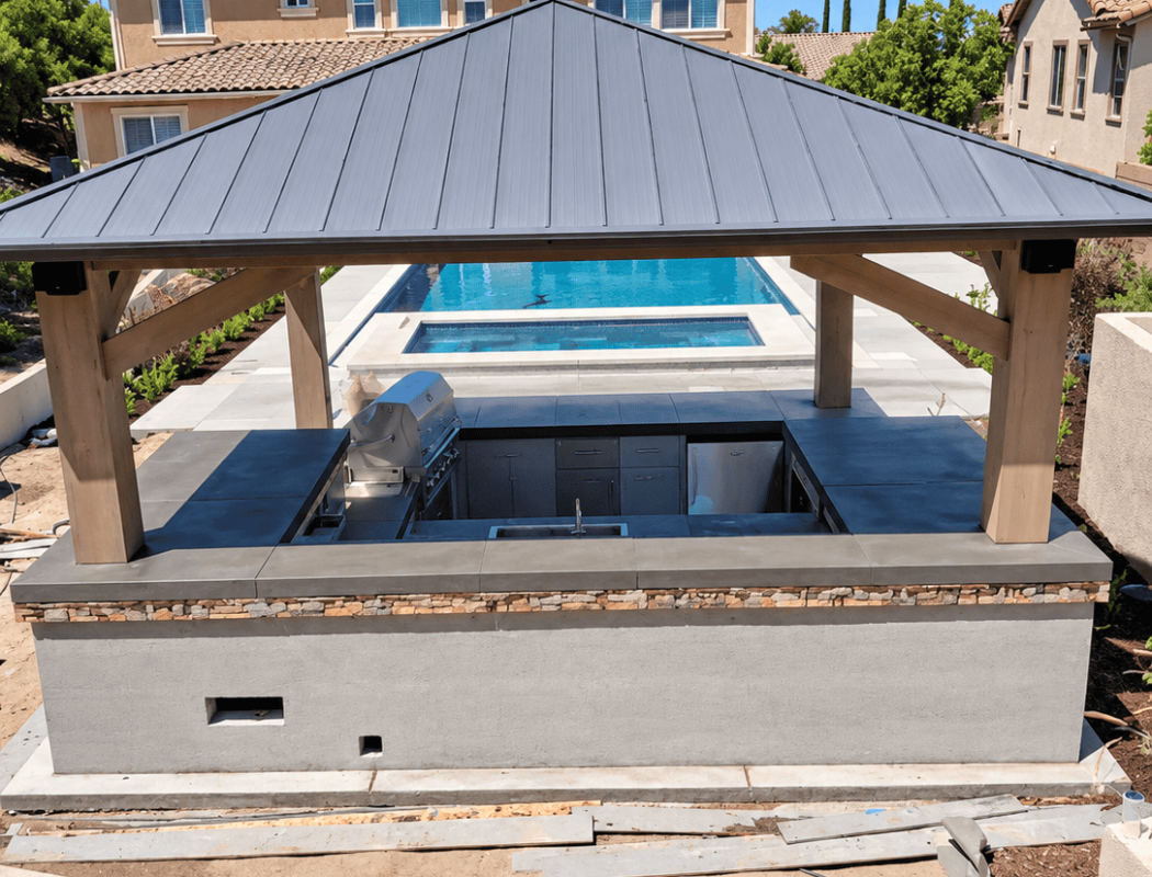 Concrete poolside barbecue area with a metal roof and built-in grill.